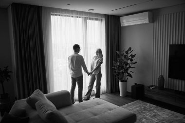 Evde pencerenin karşısında güneş ışığında duran sevimli bir çift. Yakışıklı bir adam kız arkadaşıyla el ele tutuşuyor. Sıradan giysiler içinde uzak bir çiftin görüntüsü. Siyah ve beyaz resim.