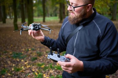 Siyah ceketli sakallı bir adam renkli bir ormanda bir dron ve kontrolör tutuyor. Heyecan verici bir uçuşa hazırlanırken parlak yapraklar etrafını sarar..