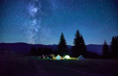 Bir grup arkadaş kamp ateşi etrafında dinleniyor, çarpıcı yıldızlı gökyüzünün altında aydınlık çadırlarla çevrili. Samanyolu uzanıyor ve huzur dolu dağlarda büyülü bir atmosfer yaratıyor..