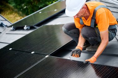 Evin çatısında fotovoltaik güneş paneli inşa eden bir işçi. Miğferlerde mühendis ve eldivenler dışarıda altıgen anahtarla güneş modülü kuruyor. Alternatif enerji üretimi kavramı.