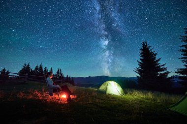 Yürüyüşçü kadın kamp ateşiyle gevşer. Ilık parıltısıyla aydınlanır. Yıldızlarla dolu gökyüzünün altında çadır kurar. Samanyolu başınızın üstüne uzanır ve dingin dağ düzenine sihirli bir dokunuş ekler..