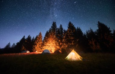Çimenli, berrak, yıldızlı gökyüzünün altında parlayan kamp ateşiyle aydınlatılmış birkaç çadır. Dağlarda yüksek çam ağaçlarıyla çevrili. Doğada kamp yaparak geçirilen gecenin huzurlu güzelliği..