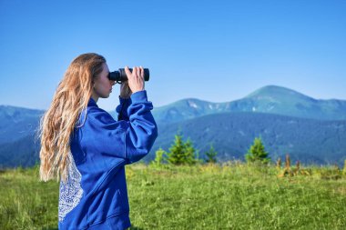 Çimenli çayırlarda duran kadın, uzak dağları gözlemlemek için dürbün kullanıyor, mavi kapüşonlu bir şey giyiyor. Sahne, yemyeşil tepelerin ve açık mavi gökyüzünün zeminine kurulur..