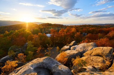 Batan güneş, sonbahar manzarası üzerine altın parıltısını saçarak canlı kırmızı ve turuncu yaprakları aydınlatıyor. Bulutlu gökyüzünün altında engebeli kaya oluşumları. Dovbush Kayaları, Karpat Dağları, Ukrayna.