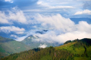 Dağ vadisinin yeşil otlaklar ve dağınık evlerle kaplanmış manzarası. Bulutlar ormanlık tepelerde yavaşça sürüklenir, sakin ve sakin bir atmosfer yaratır..