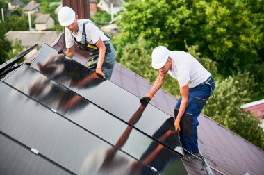 İşçiler evin çatısına güneş paneli inşa ediyorlar. Dışarıda fotovoltaik güneş modülü kuran kask takan iki adam. Alternatif, yeşil ve yenilenebilir enerji üretimi kavramı.