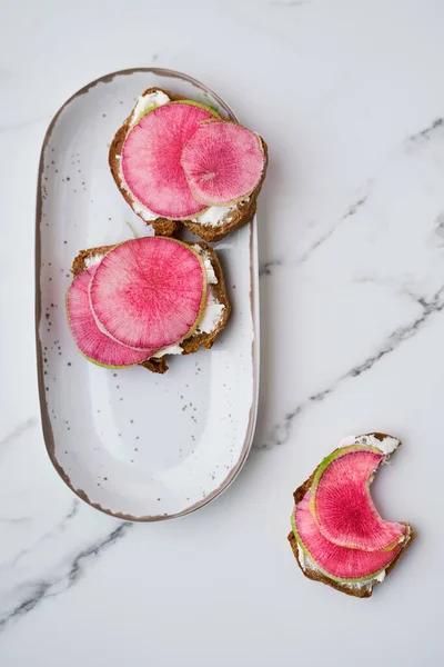Watermelon radish and cream cheese sandwiches on rye bred on oval dish, one half eaten, top view — Stock fotografie
