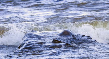 Dalgalar kıyıya vurur ve kayalara çarpar. Kıyıya yakın yerlerde bir sürü sıçrama ve sıçrama meydana getirir. Taştan çakıl taşı kıyısında dalgalı köpüklü dalgalarla fırtınalı havada nehir sörfü yapmak..