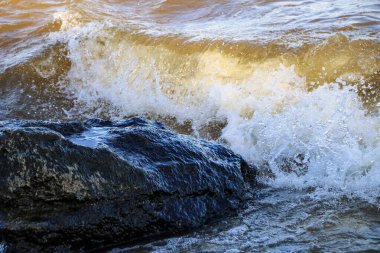 Dalgalar kıyıya vurur ve kayalara çarpar. Kıyıya yakın yerlerde bir sürü sıçrama ve sıçrama meydana getirir. Taştan çakıl taşı kıyısında dalgalı köpüklü dalgalarla fırtınalı havada nehir sörfü yapmak..