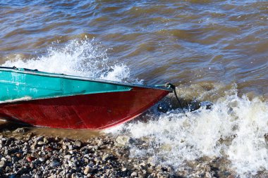 Eski paslı balıkçı teknesi nehrin çakıl taşı kıyısında uzanıyor. Dalgaların dalgaları küçük demir adamın üzerinde yuvarlanıyor ve teknenin yan tarafında köpük ve sprey haline geliyor..