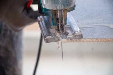 Bir inşaat işçisinin elleri elektrikli bir yapbozun parçalarını kesip biçiyor. İç dekorasyon için bir yapı tahtasını elektrikli bir yap-boz ile kesme işlemi