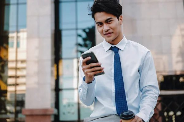 Uśmiechnięty Młody Azjatycki Biznesmen Korzystający Telefonu Komórkowego Mieście Patrząc Kamerę — Zdjęcie stockowe