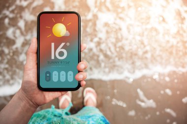 Yaz Güneşi Günü Konsepti 'nde hava güzel. Hava Durumu Hava Durumu Bilgileri 'ni görmek için Elde Tutulan Cep Telefonu. Arka planda güneş ışığı olan kumsalda hafif dalgalanma. Üst Görünüm
