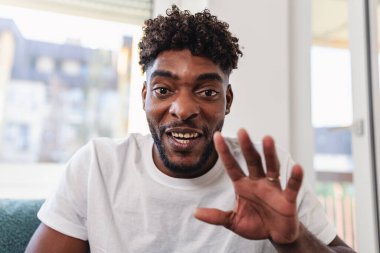 Adam evde otururken gülümsüyor ve el sallıyor.