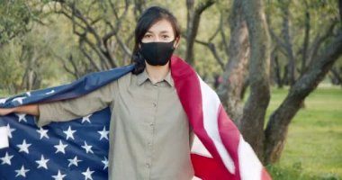 Koruyucu maskeli genç bir kadın kendini Amerikan bayrağına sarıp parktaki kameraya bakıyor. Bağımsızlık günü, seçimler ve salgın konsepti.