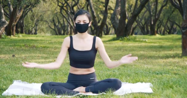 晴れた日に公園の芝生の上に座って黒い保護マスクを身に着けている間 若い女性は蓮の位置でヨガ瞑想を練習します 流行で健康的なライフスタイルのコンセプト — ストック動画
