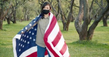 Koruyucu maskeli genç bir kadın kendini Amerikan bayrağına sarıp parktaki kameraya bakıyor. Bağımsızlık günü, seçimler ve salgın konsepti.