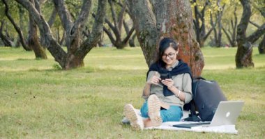 Gözlüklü genç bir kadın parkta çimlerin üzerinde otururken sosyal ağlarda haber yayını yapıyor.