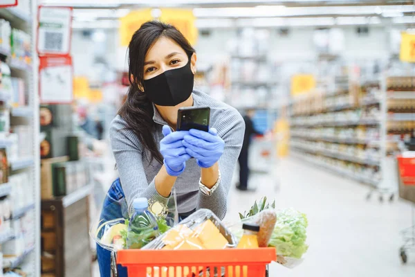 Süpermarkette alışveriş yaparken elinde akıllı telefon tutan koruyucu maskeli ve eldivenli gülümseyen genç bir kadının portresi.