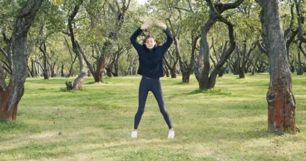 スポーツウェアを着た若い女性が公園で腕を上げてジャンプします 健康的なライフスタイルとスポーツコンセプト — ストック動画