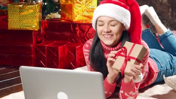 サンタ ハットの若い幸せな女性は 自宅の床に横たわっているラップトップを使用して 贈り物を与え 通信ビデオについて話しています 社会的距離 新しい通常と休日の概念 — ストック動画