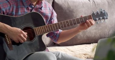 Evde dizüstü bilgisayar kullanarak akustik gitar dersi veren tanınmamış genç bir adam..
