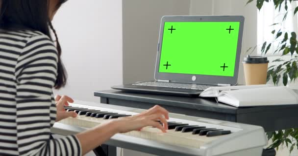 若い女性は自宅のノートパソコンを使って遠隔操作で電子ピアノを学ぶ 緑の画面だ クロマキー オンライン教育とレジャーの概念 — ストック動画