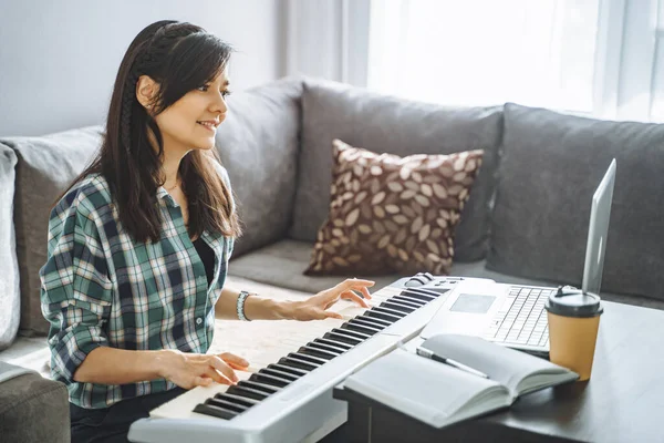 Young woman music teacher playing electric piano teaching remotely using laptop while working from home. Online education and leisure concept.