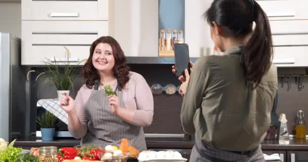Привлекательная женщина, танцующая и готовящая овощной салат, пока ее подруга снимает видео для социальных сетей на смартфоне на кухне. Концепция блогов и жизни в Интернете. — стоковое видео