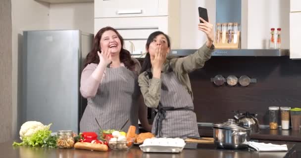 두 명의 행복 한 여성이 셀카를 먹거나 스마트폰으로 비디오를 녹화하고 부엌에서 요리하면서 춤을 추고 웃고 있습니다. 블로 깅과 소셜 미디어 개념 — 비디오