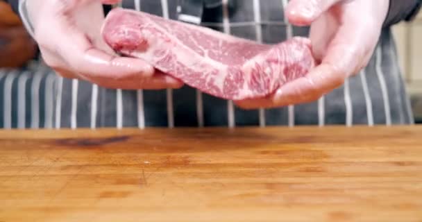 Готуємо мармуровий яловичий стейк на дерев'яній дошці. Чоловічі кухарські руки в рукавичках зморщують і кидають шматочок м'яса на стіл — стокове відео