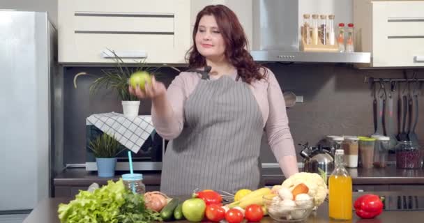 Retrato de uma mulher sorrindo plus size jogando uma maçã verde enquanto prepara um jantar saudável na cozinha. Dieta e alimentação saudável. — Vídeo de Stock