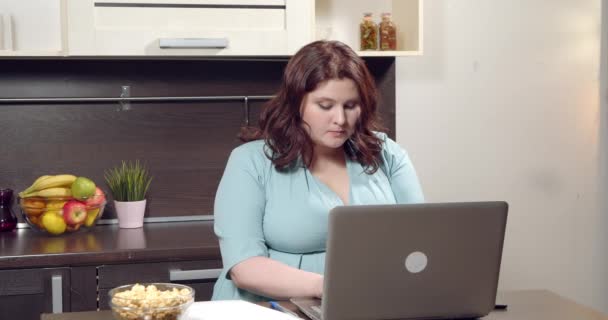 Atraente plus size jovem mulher trabalhando no laptop e comer pipoca na cozinha — Vídeo de Stock