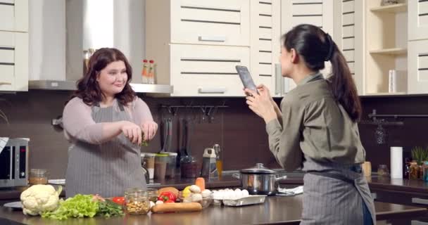Atraente plus size mulher dançando e preparando salada de legumes enquanto seu amigo está filmando vídeo para redes sociais em smartphones na cozinha. Blogging and living online concept. — Vídeo de Stock