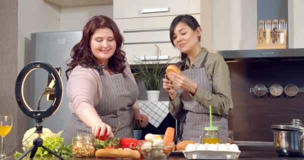 Дві веселі молоді жінки знімають відео для свого блогу або ведуть прямий ефір у соціальних мережах про створення бургерів за допомогою смартфона на кухні . — стокове відео