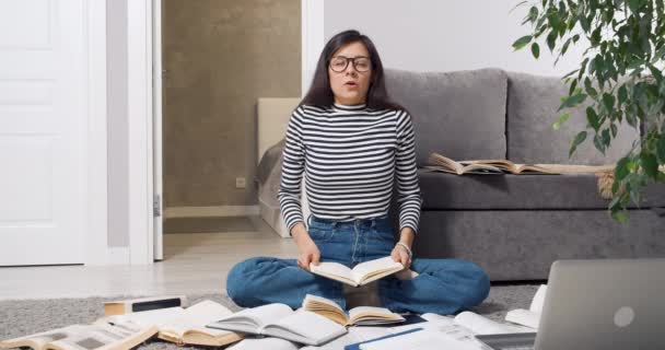 Stanco giovane studentessa con occhiali posa libro sulla testa e meditare tra mucchi di libri di testo sensazione di stress mentre si prepara per l'esame seduto sul pavimento a casa. — Video Stock