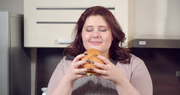 Närbild porträtt av unga plus storlek kvinna slicka och njuta av att äta läckra hamburgare i köket — Stockvideo