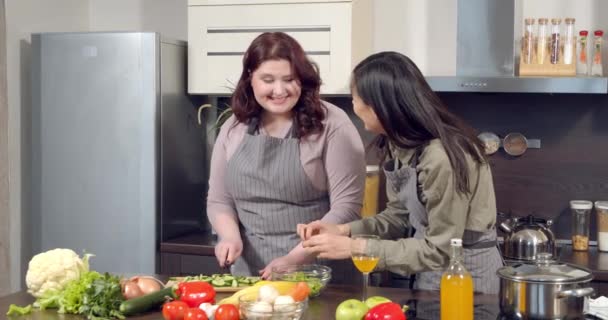 Två attraktiva unga kvinnor förbereder middag med färska grönsaker har roligt chatta och ler i köket hemma. — Stockvideo