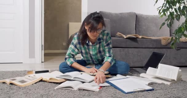 以戴眼镜的年轻女学生或女记者为焦点，坐在家里的地板上寻找许多书籍和教科书中的信息。教育概念 — 图库视频影像