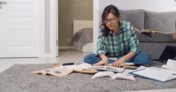 O tânără studentă sau jurnalistă concentrată în ochelari care caută informații în multe cărți și manuale în timp ce stătea pe podea acasă. Conceptul de educație — Videoclip de stoc