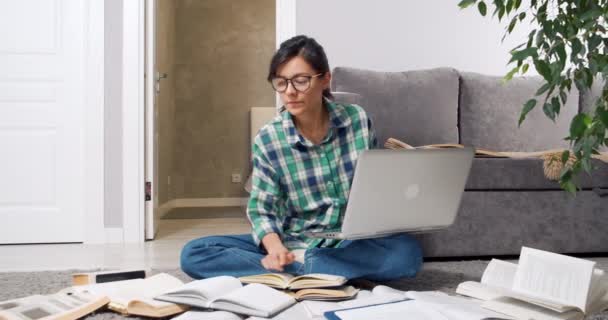 Estudiante o periodista joven concentrada con gafas buscando información en muchos libros y libros de texto y escribiendo algo en una computadora portátil en casa. Concepto educativo — Vídeos de Stock