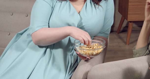 Неузнаваемая плюс женщина размером с попкорн, сидящая на диване. Руки женщин, принимающих закуски из миски во время просмотра фильма дома. — стоковое видео