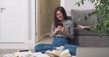 Gözlüklü çekici bir kadın akıllı telefon kullanarak iletişim kurarken gülümsüyor ve evde oturmuş ders kitapları ve kitapları öğrenmeye devam ederken iç çekiyor.
