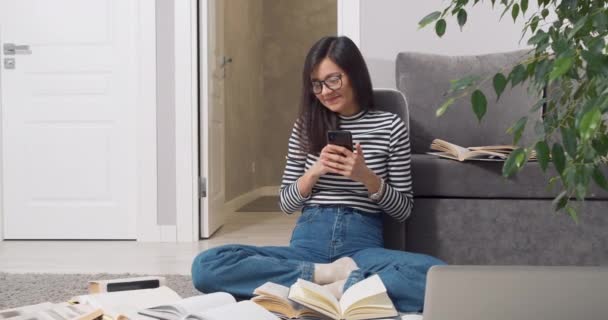 Attraktive Frau mit Brille, die lächelt, während sie online mit einem Smartphone kommuniziert und heftig seufzt, während sie zu Hause auf dem Boden sitzend Lehrbücher und Bücher lernt — Stockvideo