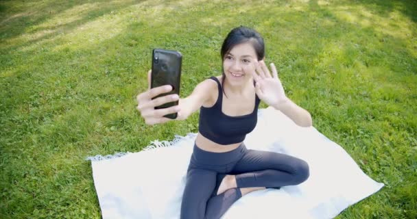 スポーツウェアの録画ビデオで若い魅力的な女性や公園内の芝生の上に座ってスマートフォンを使用して友人とオンライン会議を持っている。ブログ、ソーシャルメディア、オンラインコミュニケーション — ストック動画
