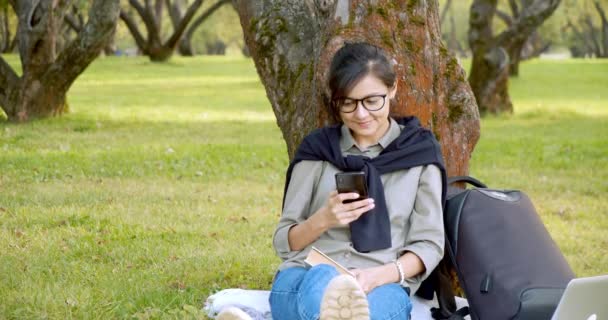 Ung leende kvinna i glasögon rulla en nyhetssändning i sociala nätverk sitter på gräset i parken. — Stockvideo