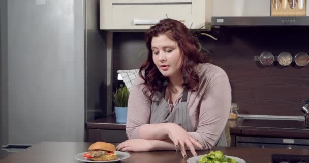 Молодая женщина плюс размер трудно выбрать между зеленым салатом и бургером и улыбаясь, принимая тарелку с вкусным чизбургером на кухне. — стоковое видео