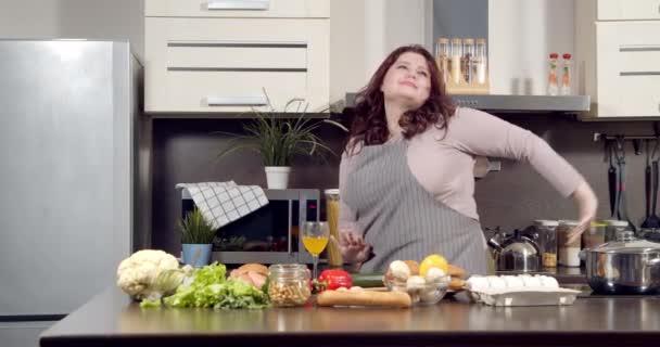 Vonzó plusz méret fiatal nő táncol energikus főzés közben a konyhában. A test pozitív koncepciója — Stock videók