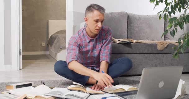 Koncentrerad ung man söker information i många böcker och läroböcker och skriva något i en bärbar dator hemma. Koncept för utbildning och distansarbete. — Stockvideo
