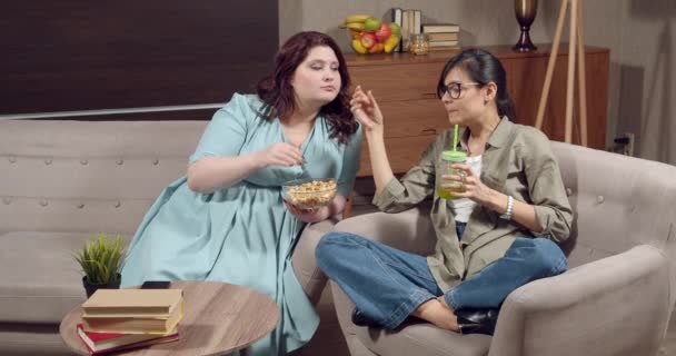 Dos amigas comiendo palomitas de maíz mientras miran la televisión y discuten una película sentadas en la sala de estar en casa — Vídeo de stock